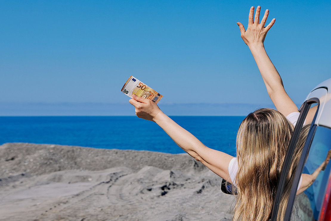 Happy woman leaning out of car window holding 50 euro banknotes and waving at the sea. Concept of travel budget, vacation freedom, and wealth.