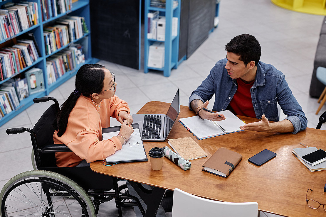 Asian teenage girl with disability talking to friend in college library
