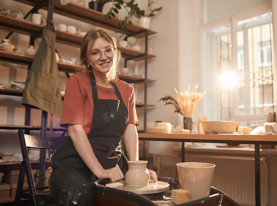 Warm toned portrait of young female artisan smiling at camera while working on pottery wheel in sunlit workshop and enjoying arts and crafts, copy space