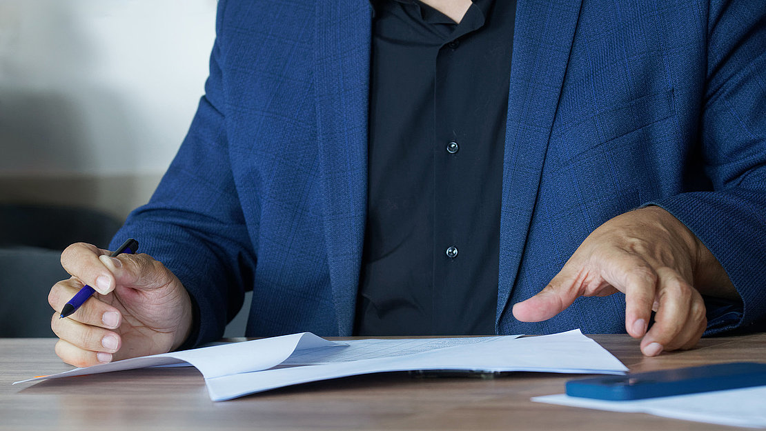An adult man in a business suit, sitting at a table with a pen, fills out an agreement or order form. Concept of concluding an agreement or filling out forms. Photo. No face. Selective focus