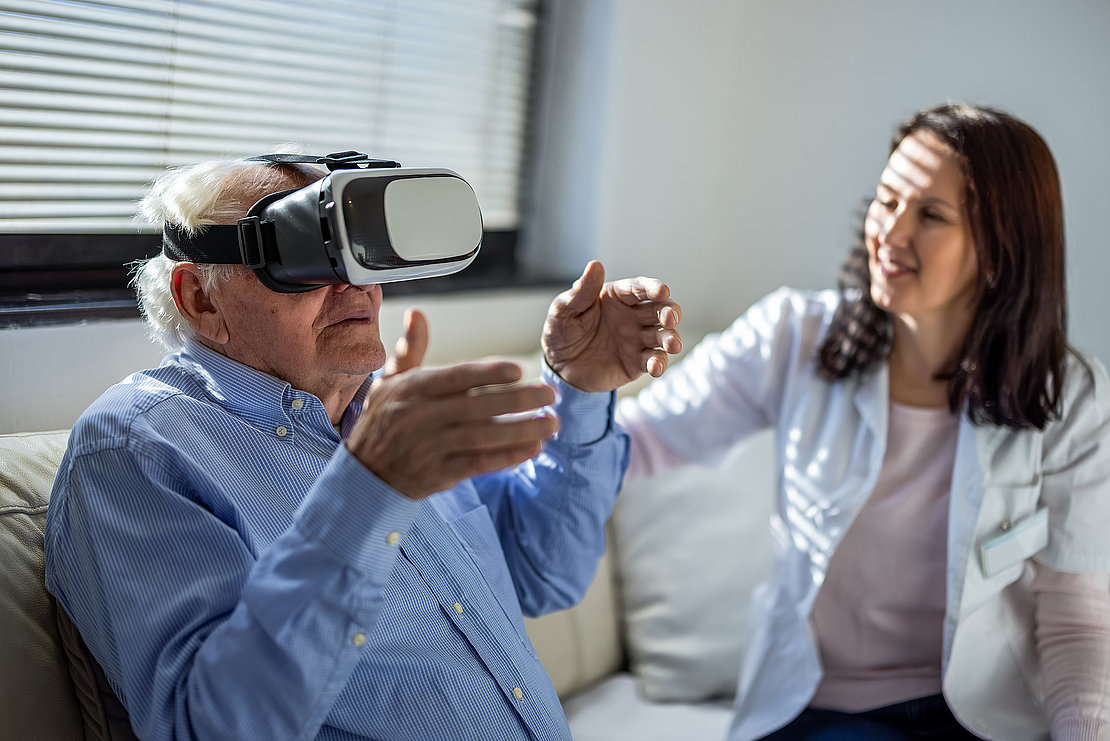 Senior man using a virtual reality headset while a nurse sitting in the background at a nursing home.