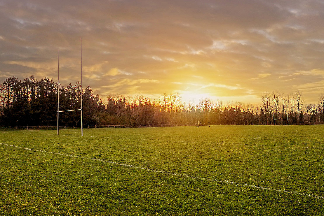 Irish National sport camogie, hurling, football and rugby tall goalposts and training ground at sunset. Selective focus, Sun flare. Man walking dog in the background out of focus.