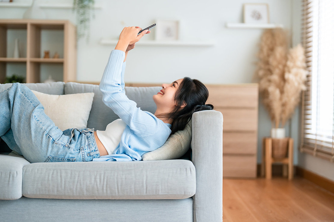 Happy cheerful positive young Asian woman on the phone at home, smiling girl using app and browsing on the internet on smartphone lying on sofa